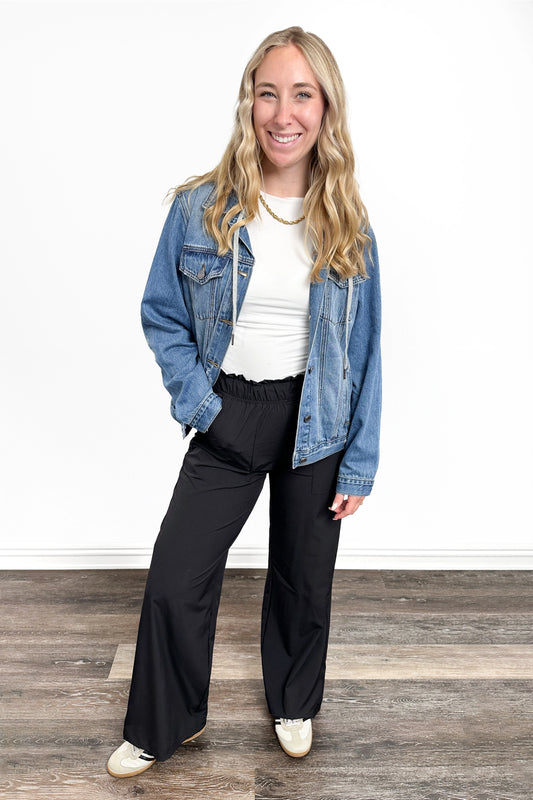 Woman wearing a blue denim jacket, white shirt, and black pants on a wooden floor.