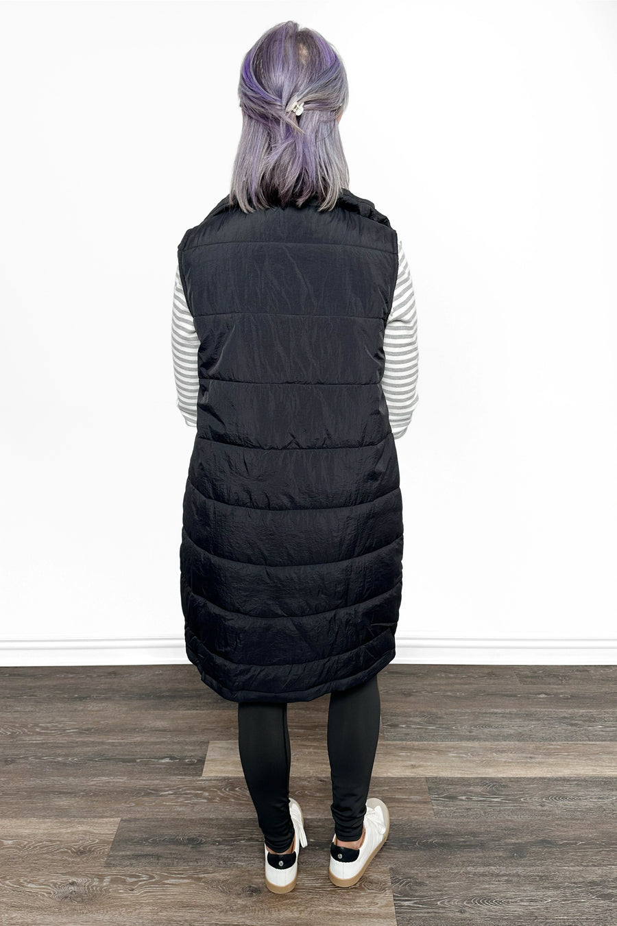 Person wearing a long black puffer vest on a wooden floor with a white wall background