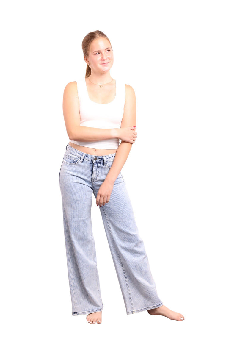 A young woman with blonde hair tied back, wearing a white ribbed tank top and light-wash wide-leg denim jeans, standing against a solid white background.