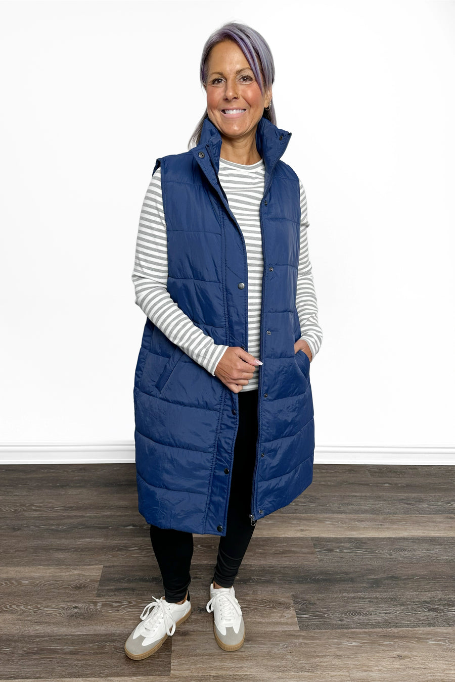 Woman wearing a long blue puffer vest over a striped shirt on a wooden floor.