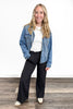 Woman wearing a blue denim jacket, white shirt, and black pants on a wooden floor.
