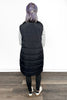 Person wearing a long black puffer vest on a wooden floor with a white wall background