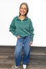 Woman wearing a green fleece pullover and blue jeans standing on a wooden floor.
